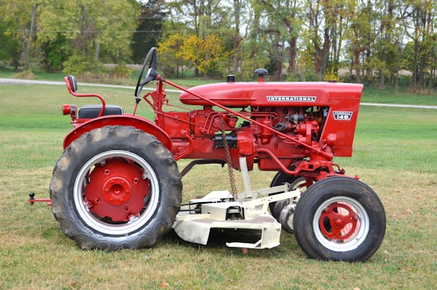 A red vintage tractor is parked on a grassy field. The tractor has large rear wheels and smaller front wheels. It features detailed mechanical components and metal parts, reflecting its classic design. The background shows a line of trees with green and yellow foliage, suggesting a rural setting.