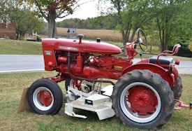 A vintage red tractor, specifically an International Harvester Farmall 140, is parked on a grassy area next to a road. The tractor is in good condition, with visible wheels, a steering wheel, and a mower attachment underneath. In the background, there are green trees, a small wooden building, and a cornfield, suggesting a rural setting.