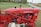 A vintage red tractor with the label 'International Farmall 140' displayed prominently. The machinery is detailed, with visible red metal components and various mechanical parts. It is situated on a patch of green grass, with a rural road and fields in the background.