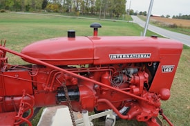 A vintage red tractor with the label 'International Farmall 140' displayed prominently. The machinery is detailed, with visible red metal components and various mechanical parts. It is situated on a patch of green grass, with a rural road and fields in the background.