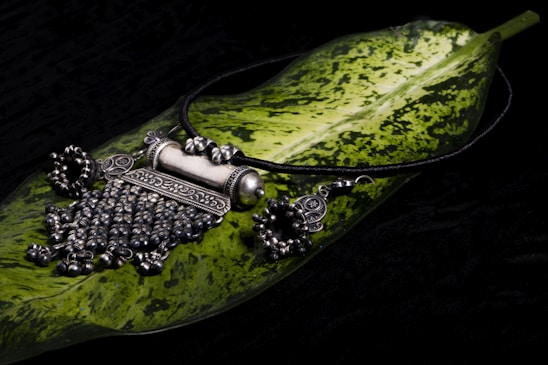 A piece of intricately designed silver jewelry, possibly a necklace, is placed on a large green leaf with variegated patterns. The jewelry features detailed craftsmanship with ornate elements and dangling beads, set against a dark background.