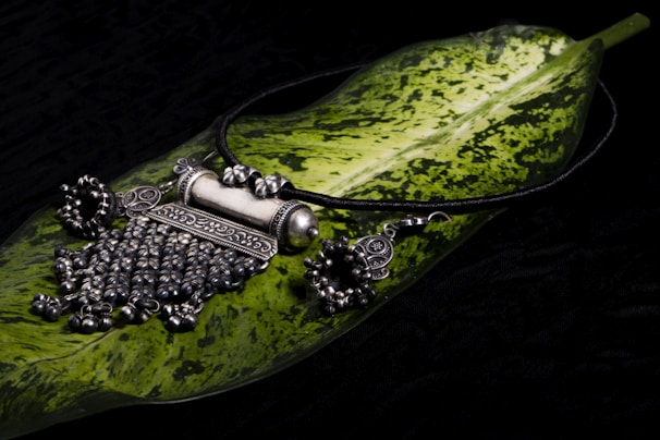 A piece of intricately designed silver jewelry, possibly a necklace, is placed on a large green leaf with variegated patterns. The jewelry features detailed craftsmanship with ornate elements and dangling beads, set against a dark background.