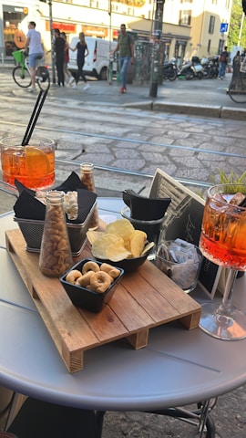 Outdoor café table set with a wooden serving tray holding various snacks including nuts, chips, and small bread rings. Two glasses of an orange-colored beverage with ice and straws are placed on the table. Behind the table, there is a street scene with pedestrians walking, a person riding a bicycle, parked motorcycles, and buildings in the background.