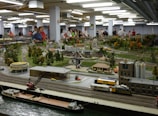 A large indoor model train exhibit depicts a detailed miniature landscape. Various model train tracks and trains are integrated with detailed miniature buildings, roads, and trees. Numerous people are gathered around the exhibit, observing and taking pictures. The setup includes different areas representing urban, suburban, and industrial zones.