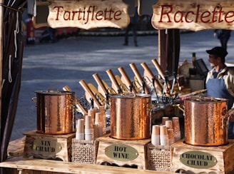A market stall features copper pots labeled 'Vin Chaud', 'Hot Wine', and 'Chocolat Chaud'. Wooden signs overhead display the names 'Tartiflette' and 'Raclette'. Stacks of paper cups are placed near the pots, and a person wearing a cap and apron stands nearby, possibly attending the stall.