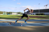 A person is sprinting on a track with determination, captured in mid-stride. The athlete is wearing a black and white striped athletic outfit and bright blue running shoes. Nearby, a stadium with a field and a large building can be seen under a clear blue sky.