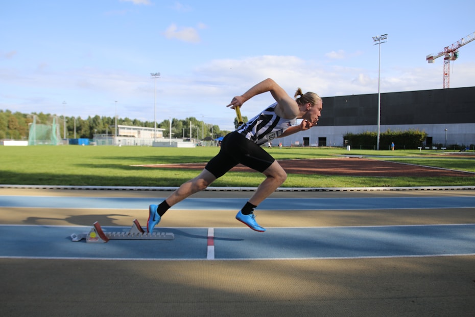 A person is sprinting on a track with determination, captured in mid-stride. The athlete is wearing a black and white striped athletic outfit and bright blue running shoes. Nearby, a stadium with a field and a large building can be seen under a clear blue sky.