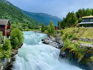 A picturesque landscape featuring a rushing river cascading down rocks amidst lush greenery. Traditional wooden houses are nestled alongside the river, surrounded by dense forests and mountains under a bright sky. The scenery conveys tranquility and a connection to nature.