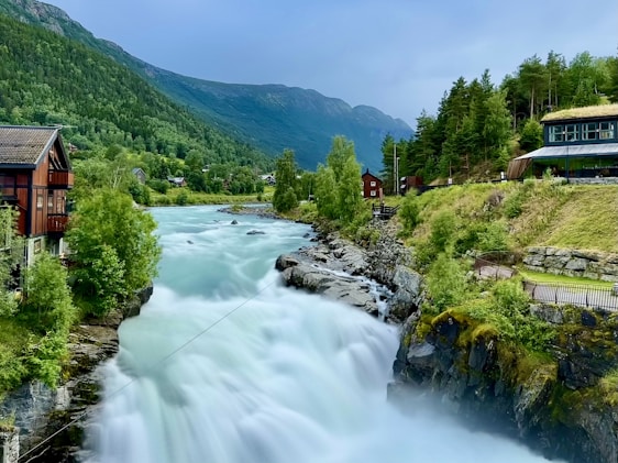 A picturesque landscape featuring a rushing river cascading down rocks amidst lush greenery. Traditional wooden houses are nestled alongside the river, surrounded by dense forests and mountains under a bright sky. The scenery conveys tranquility and a connection to nature.