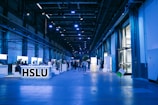 An indoor exhibition hall with high ceilings lit in blue lighting. Several informational booths line the sides, with a prominent booth labeled 'HSLU' in the foreground. People are walking and interacting throughout the space, creating a lively atmosphere.