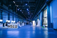 An indoor exhibition hall with high ceilings lit in blue lighting. Several informational booths line the sides, with a prominent booth labeled 'HSLU' in the foreground. People are walking and interacting throughout the space, creating a lively atmosphere.