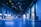 An indoor exhibition hall with high ceilings lit in blue lighting. Several informational booths line the sides, with a prominent booth labeled 'HSLU' in the foreground. People are walking and interacting throughout the space, creating a lively atmosphere.
