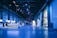 An indoor exhibition hall with high ceilings lit in blue lighting. Several informational booths line the sides, with a prominent booth labeled 'HSLU' in the foreground. People are walking and interacting throughout the space, creating a lively atmosphere.