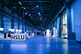 An indoor exhibition hall with high ceilings lit in blue lighting. Several informational booths line the sides, with a prominent booth labeled 'HSLU' in the foreground. People are walking and interacting throughout the space, creating a lively atmosphere.