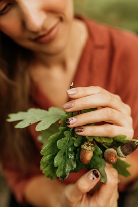 A person gently holds a small branch with vibrant green oak leaves and acorns. The focus is on the hands, which have neatly manicured nails with artistic designs. The individual's shirt is a warm, earthy tone, and the background is softly blurred, emphasizing the natural elements.
