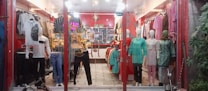 A clothing store display with mannequins dressed in various outfits. The store features brightly colored garments including shirts, dresses, and pants, arranged neatly. The interior is warmly lit, with red walls and decorative lighting fixtures on the ceiling. Shelves and racks are lined with additional clothing items.