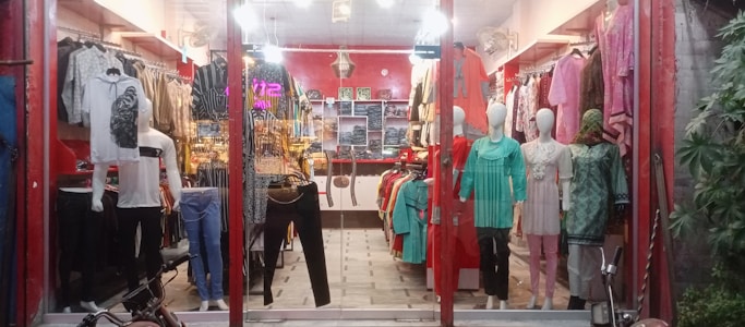 A clothing store display with mannequins dressed in various outfits. The store features brightly colored garments including shirts, dresses, and pants, arranged neatly. The interior is warmly lit, with red walls and decorative lighting fixtures on the ceiling. Shelves and racks are lined with additional clothing items.