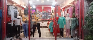 A clothing store display with mannequins dressed in various outfits. The store features brightly colored garments including shirts, dresses, and pants, arranged neatly. The interior is warmly lit, with red walls and decorative lighting fixtures on the ceiling. Shelves and racks are lined with additional clothing items.