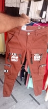 A pair of brown pants with multiple pockets and zippers is being held up by a hand. The pants are decorated with text that reads 'FUN TODAY' and 'REAL DEAL'. The background includes various hanging clothing items in different colors.