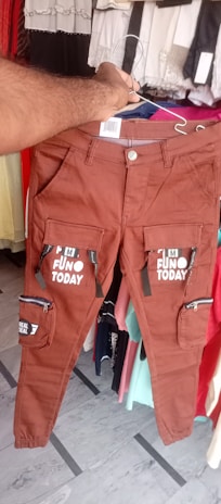 A pair of brown pants with multiple pockets and zippers is being held up by a hand. The pants are decorated with text that reads 'FUN TODAY' and 'REAL DEAL'. The background includes various hanging clothing items in different colors.