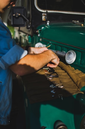 A person wearing a blue shirt is working on a green vehicle, possibly performing maintenance or repairs. Several tools are laid out on a fabric surface on top of the hood, and the individual is focused intently on his task.