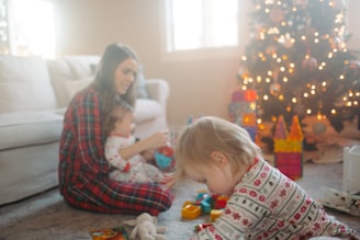 A cozy setting with a woman and two children sitting close together on a carpeted floor. They are wearing festive pajamas, and there are toys scattered around them. In the background, a decorated Christmas tree is lit with warm string lights, adding a festive atmosphere.