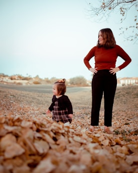 A young child in a patterned dress and dark sweater stands amidst fallen autumn leaves. Next to the child, an adult woman in a rust-colored top and black pants looks on with hands on hips. The background features a light blue sky and a bare tree branch protruding from the top right corner.