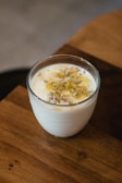 A clear glass filled with creamy yogurt topped with a drizzle of honey and sprinkled with a small amount of seeds. The glass is placed on a wooden surface with a blurred background, suggesting a cozy, warm setting.