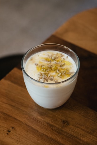 A clear glass filled with creamy yogurt topped with a drizzle of honey and sprinkled with a small amount of seeds. The glass is placed on a wooden surface with a blurred background, suggesting a cozy, warm setting.