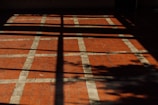 A floor with a pattern of square tiles, where the tiles are primarily red with white grid lines. The floor is partially covered by shadows, which create a cross-like pattern and the silhouette of foliage.