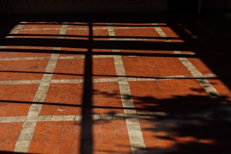 A floor with a pattern of square tiles, where the tiles are primarily red with white grid lines. The floor is partially covered by shadows, which create a cross-like pattern and the silhouette of foliage.