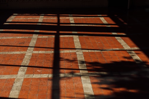 A floor with a pattern of square tiles, where the tiles are primarily red with white grid lines. The floor is partially covered by shadows, which create a cross-like pattern and the silhouette of foliage.