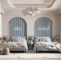 Two twin beds with light blue upholstered headboards are positioned against a wall with arched niches. Above each bed, there are names lit up in neon lights. Between the beds, a small table holds a vase with flowers. Plush toys are on the beds and a children's wooden toy whale is on the floor in front of one of the beds. A cloud-shaped chandelier hangs from the ceiling, and soft, fluffy blankets and pillows adorn the beds. The room's decor includes soft, neutral tones with hints of light blue.
