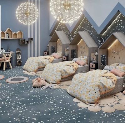 Three children's beds with house-shaped headboards are placed against a wall decorated with mountain peaks and star patterns. The room is illuminated by two spherical chandeliers with a starry appearance, creating a whimsical atmosphere. A small table with stools and various decor, such as a skateboard and toy house shelves, are visible. The bedding features polka dot patterns in soft pastel colors, complementing the star-patterned carpet.