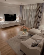A cozy living room with modern decor includes a large flat-screen television mounted on the wall above a white media console. Two lamps with beige shades are placed on either end of the console, emitting a warm glow. An armchair with a pillow and a socket is positioned next to floor-to-ceiling curtains with twinkling fairy lights behind them. A beige sectional sofa adorned with patterned throw pillows is paired with a marble-top coffee table. On the table, there is a vase of pink tulips and a few lit candles. A rug lies under the coffee table, and the room features wooden flooring.