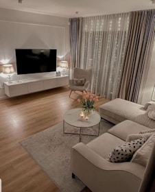 A cozy living room with modern decor includes a large flat-screen television mounted on the wall above a white media console. Two lamps with beige shades are placed on either end of the console, emitting a warm glow. An armchair with a pillow and a socket is positioned next to floor-to-ceiling curtains with twinkling fairy lights behind them. A beige sectional sofa adorned with patterned throw pillows is paired with a marble-top coffee table. On the table, there is a vase of pink tulips and a few lit candles. A rug lies under the coffee table, and the room features wooden flooring.
