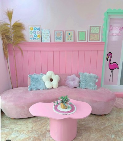 A cozy room with a pastel pink theme, featuring a curved pink sofa adorned with decorative cushions shaped like flowers. Above the sofa, a series of framed artworks line the wall, displaying cheerful designs and inspirational text. The room includes a door labeled 'flamingo room' with an illustration of a flamingo. A small pink coffee table holds a plant in a decorative pot.