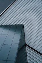 An abstract architectural design featuring intersecting lines of a modern building. The structure is composed of sleek, reflective glass panels and diagonally aligned metallic siding, creating a striking geometric pattern.