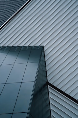 An abstract architectural design featuring intersecting lines of a modern building. The structure is composed of sleek, reflective glass panels and diagonally aligned metallic siding, creating a striking geometric pattern.