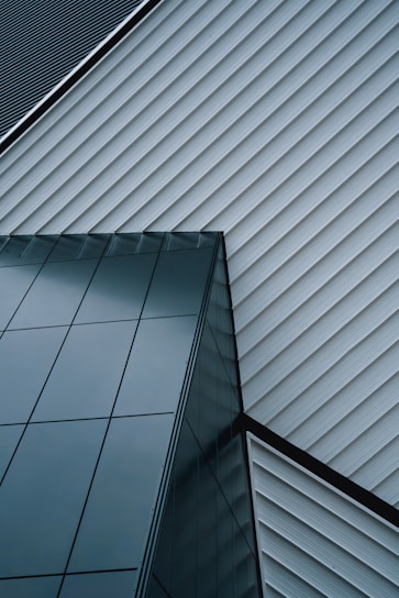 An abstract architectural design featuring intersecting lines of a modern building. The structure is composed of sleek, reflective glass panels and diagonally aligned metallic siding, creating a striking geometric pattern.