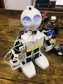 A robot with a humanoid shape and bright blue lights for eyes stands on a wooden surface. It is mainly white with black accents and is connected with various wires. Several small robotic vehicles with electronic components are visible in the background.