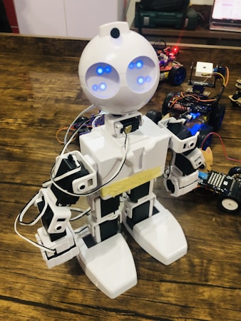 A robot with a humanoid shape and bright blue lights for eyes stands on a wooden surface. It is mainly white with black accents and is connected with various wires. Several small robotic vehicles with electronic components are visible in the background.