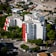 Two tall, modern residential buildings with white facades and bold red accents are surrounded by lush green trees. The area is structured with roads that have parked cars and visible pedestrian crossings, while smaller buildings and industrial structures can be seen in the background.