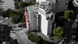 An aerial view of a modern, multi-story residential building with a mostly white facade and some red accents. Surrounding the building are several lush green trees, and a parking area with numerous parked cars. The building's rooftop features solar panels and other equipment.