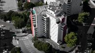 An aerial view of a modern, multi-story residential building with a mostly white facade and some red accents. Surrounding the building are several lush green trees, and a parking area with numerous parked cars. The building's rooftop features solar panels and other equipment.