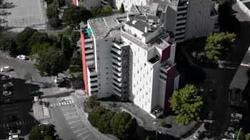 An aerial view of a modern, multi-story residential building with a mostly white facade and some red accents. Surrounding the building are several lush green trees, and a parking area with numerous parked cars. The building's rooftop features solar panels and other equipment.