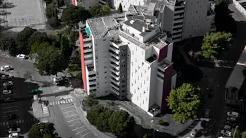 An aerial view of a modern, multi-story residential building with a mostly white facade and some red accents. Surrounding the building are several lush green trees, and a parking area with numerous parked cars. The building's rooftop features solar panels and other equipment.