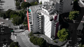 An aerial view of a modern, multi-story residential building with a mostly white facade and some red accents. Surrounding the building are several lush green trees, and a parking area with numerous parked cars. The building's rooftop features solar panels and other equipment.