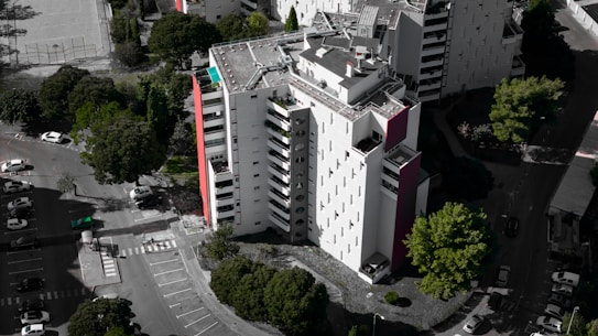 An aerial view of a modern, multi-story residential building with a mostly white facade and some red accents. Surrounding the building are several lush green trees, and a parking area with numerous parked cars. The building's rooftop features solar panels and other equipment.