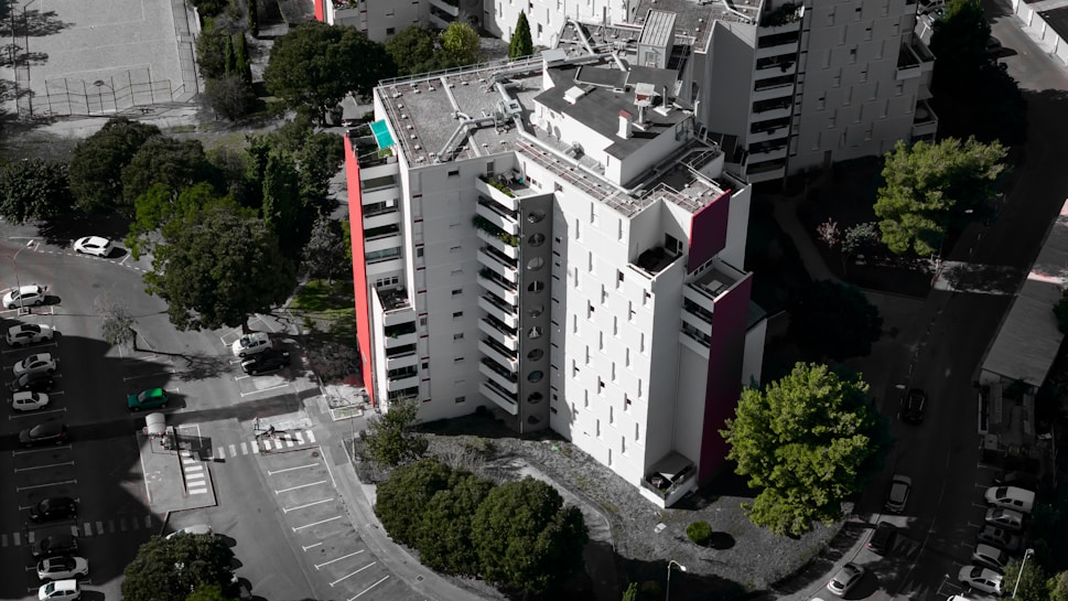 An aerial view of a modern, multi-story residential building with a mostly white facade and some red accents. Surrounding the building are several lush green trees, and a parking area with numerous parked cars. The building's rooftop features solar panels and other equipment.